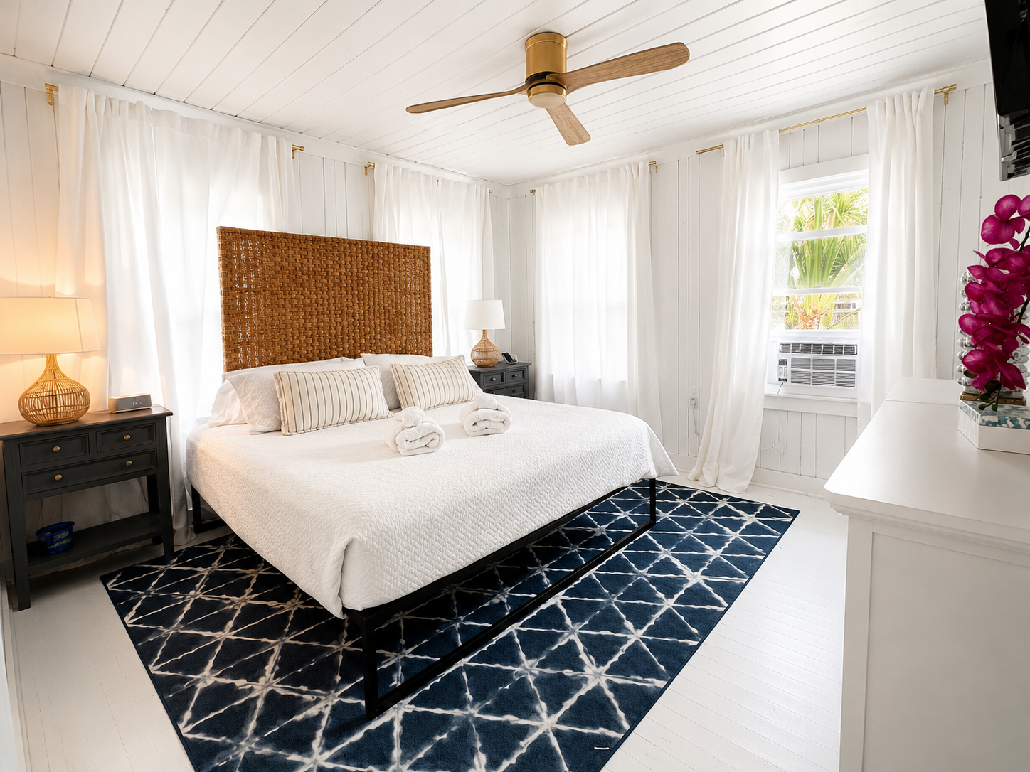 A sophisticated screened back porch at The Blue Anchor with teak lounge chairs, an ivory cushion, and glimpses of palmettos and a sandy lane just beyond.
